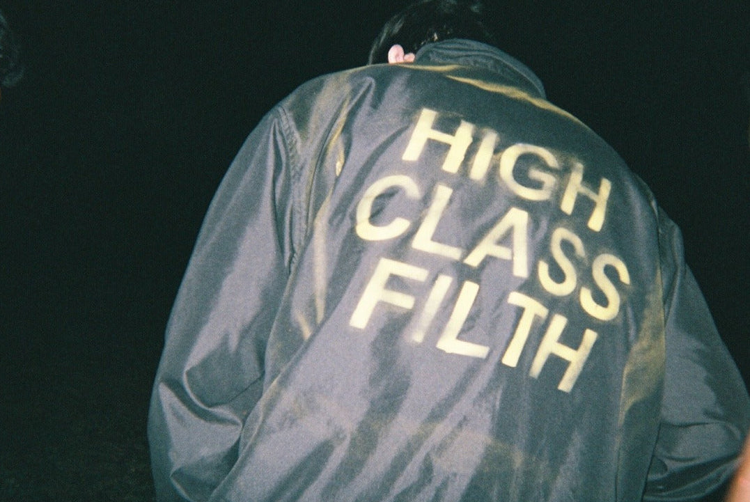 Person wearing a jacket with 'HIGH CLASS FILTH' text in a dark setting